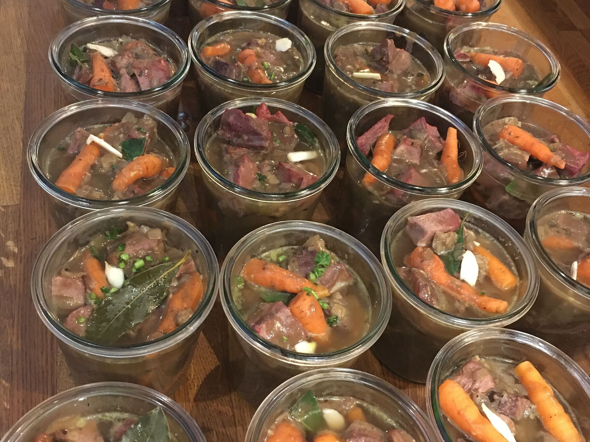 Multiple clear glasses of soup with visible ingredients like carrots, meat, and herbs.