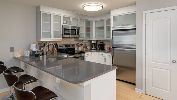 Una cocina moderna con armarios blancos, electrodomésticos de acero inoxidable, una isla central con taburetes y una puerta a la derecha.
