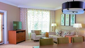 Ein Hotelzimmer mit Fernseher, einer Couch, einem Esstisch und einem großen Fenster mit Vorhängen.