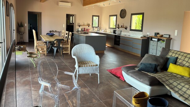 A modern kitchen with a central island, dining area with chairs, and a living room with a sofa and cushions.