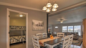 A dining area with a dark wooden table, white chairs, and a chandelier. There is a painting on the wall and a window with blinds in the background.