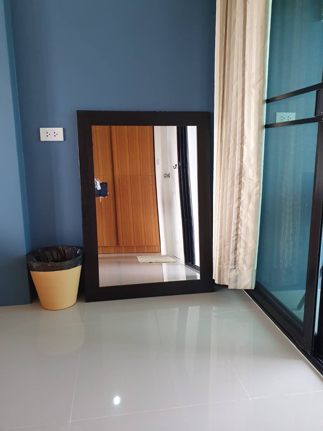 A room with a large mirror reflecting a door, a trash bin, and a window with curtains.