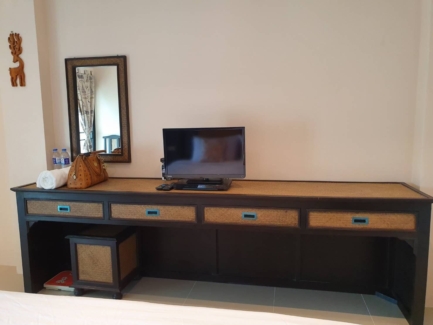 Une chambre d’hôtel avec un bureau en bois foncé, un téléviseur à écran plat, un miroir et une petite étagère sur laquelle se trouvent une télécommande et une bouteille.