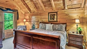 Een slaapkamer in een houten chalet met een groot bed, twee nachtkastjes, een stoel en een raam met gordijnen.