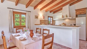A dining area with a table set for four, wooden chairs, a kitchen with a white refrigerator, and a window with curtains. - Villa Joana with Mountain View, Wi-Fi, Garden, Pool & Terraces (Pollensa)
