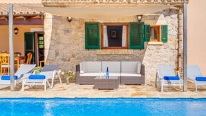 A poolside area with a stone wall, green shutters, a white sofa, and lounge chairs. - Villa Joana with Mountain View, Wi-Fi, Garden, Pool & Terraces (Pollensa)