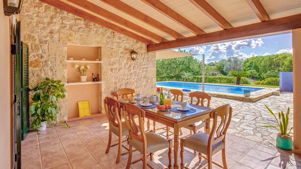 A covered patio with a stone wall, wooden ceiling beams, a dining table with chairs, a pool, and potted plants. - Villa Joana with Mountain View, Wi-Fi, Garden, Pool & Terraces (Pollensa)