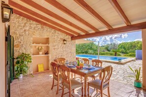 Un patio cubierto con pared de piedra, vigas de madera en el techo, una mesa de comedor con sillas, una piscina y plantas en macetas.