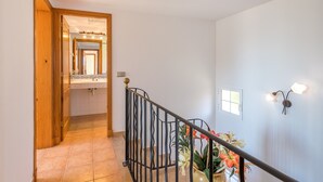 Un corridoio con una porta in legno, un bagno con uno specchio, un pavimento piastrellato, una ringhiera in metallo nero e un lampadario a parete.