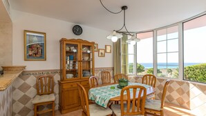 Zona pranzo con tavolo apparecchiato per quattro, circondato da sedie, un mobile in legno con stoviglie e vista mare attraverso ampie finestre.