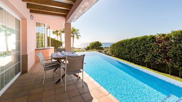 Zona piscina con tavolo e sedie, vista sul mare e siepe curata.