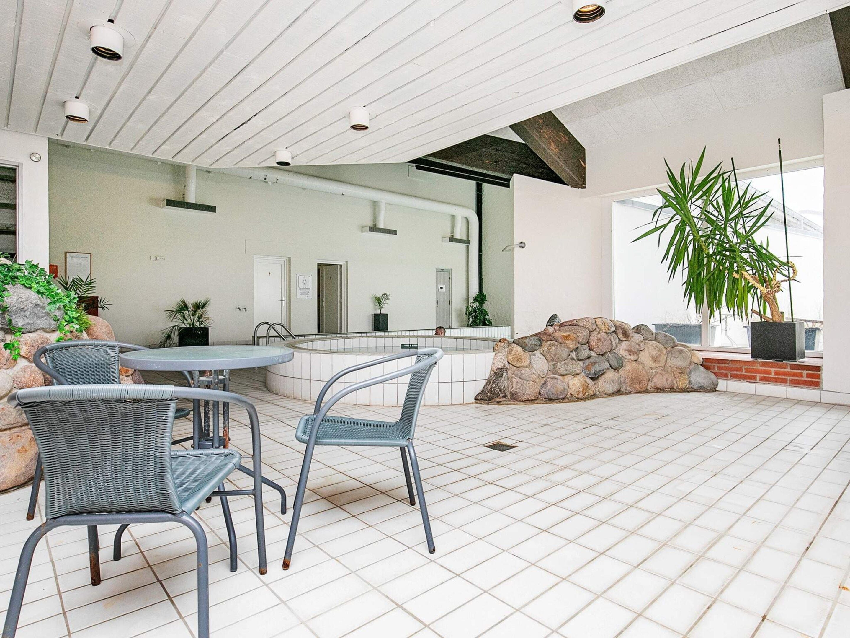 A modern indoor pool area with stone walls, a seating area with chairs, and potted plants.