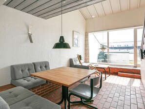 A room with a wooden table, grey upholstered seating, a green pendant light, and a view through a large window.