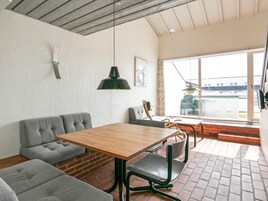 Una habitación con una mesa de madera, asientos tapizados en gris, una lámpara colgante verde y vistas a través de un ventanal grande.