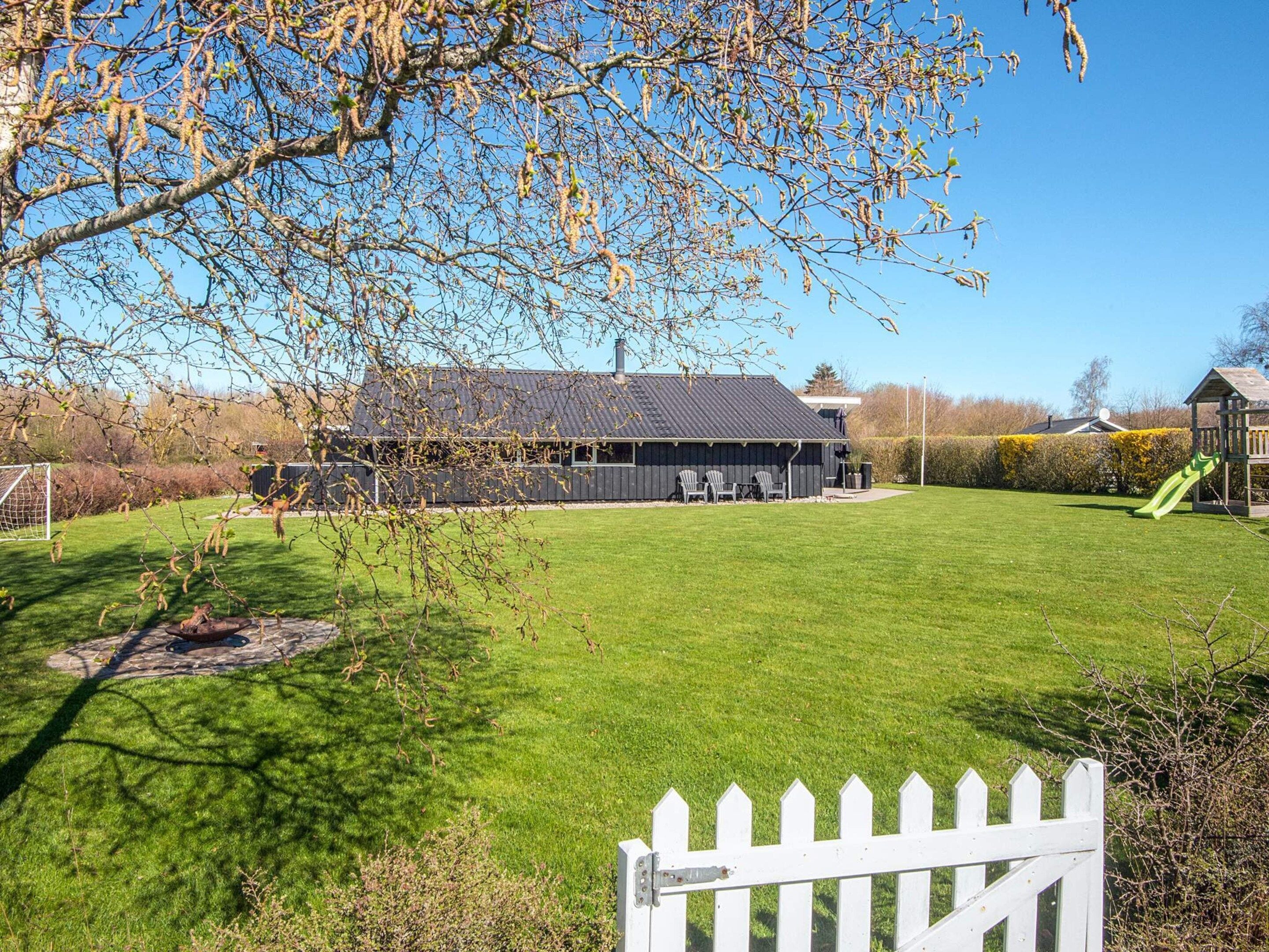 A grassy yard with a white picket fence, a building with a dark roof, and a playground with a slide.