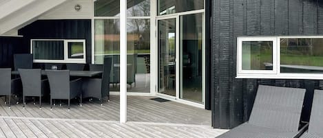 A modern outdoor deck with a dining area, large sliding glass doors, and a dark wooden wall.