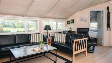 En moderne stue med en sort lædersofa, en træstol og et sofabord. Der er vinduer der lukker naturligt lys ind og giver udsigt til grønne omgivelser.
