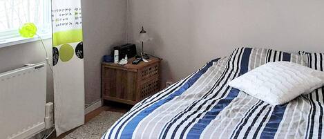 Una habitación con cama, mesita de noche, radiador y ventana con cortinas.