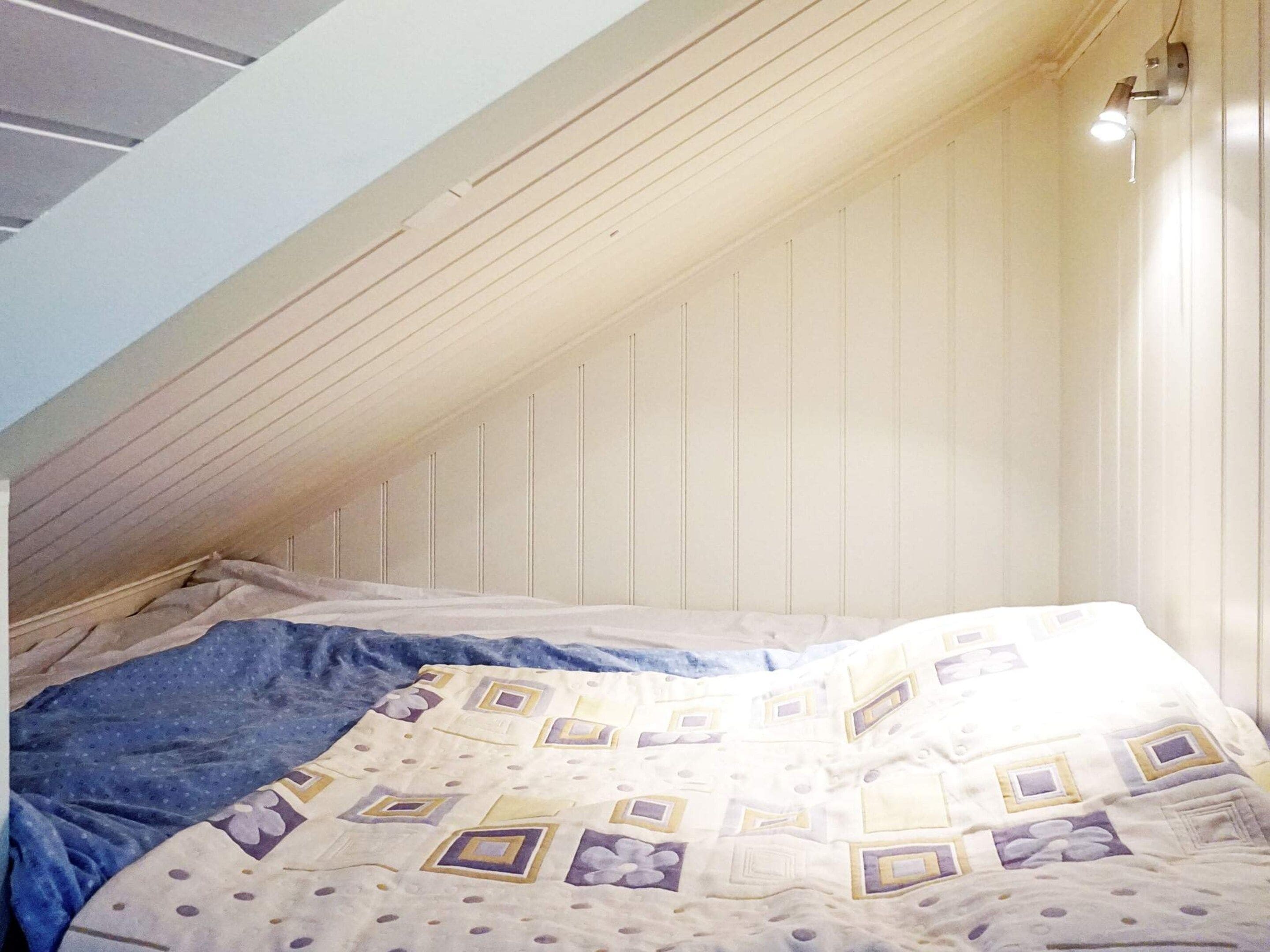 Una habitación con techo inclinado, una cama con colcha estampada y una lámpara fijada a la pared.