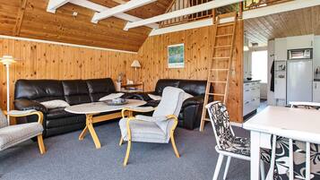 A cozy interior with a black leather sofa, a small round table, and a white dining table with chairs. A wooden ladder and a white refrigerator are visible in the background.