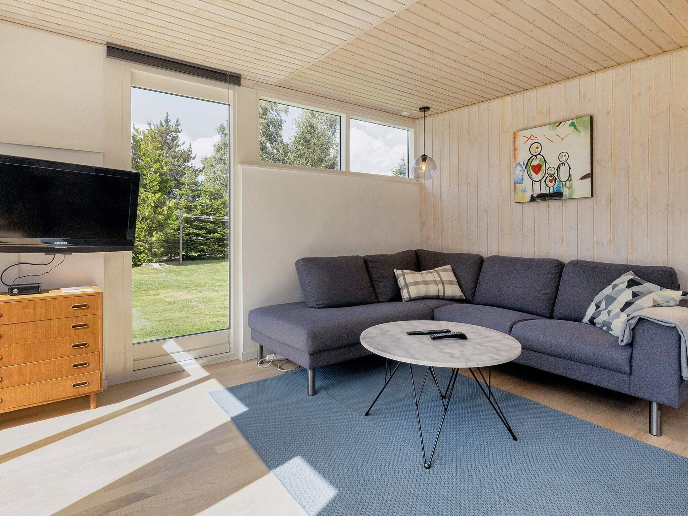 A modern living room with a grey sectional sofa, a round coffee table, a flat-screen TV mounted on the wall, and a wooden cabinet with drawers.