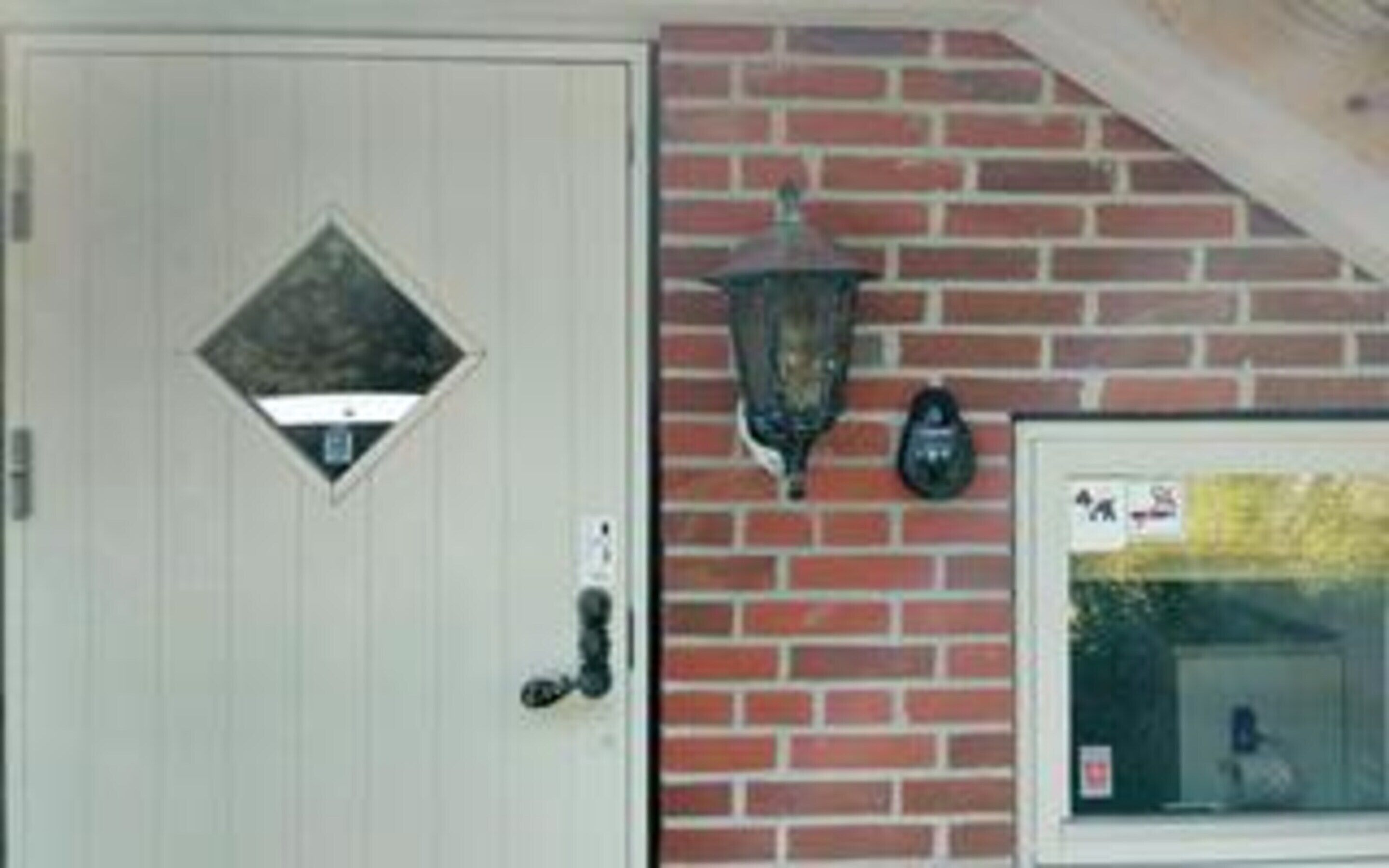 A white door with a diamond-shaped window, a black wall-mounted lantern, and a window with a view of a tree.