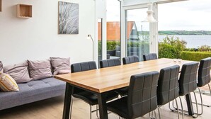Een moderne eethoek met een houten tafel en zwarte stoelen, een grijze bank met kussens, en een groot raam met uitzicht op een wateroppervlak.