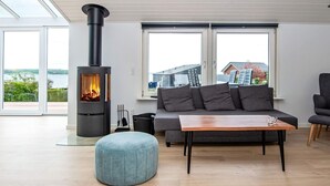 Een moderne woonkamer met een houtkachel, een grijze bank, een houten salontafel en grote ramen met uitzicht op een waterpartij.