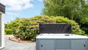 A hot tub with steps and a cover, surrounded by lush greenery and a paved patio. - 8 Person Holiday Home in Henne (Henne)