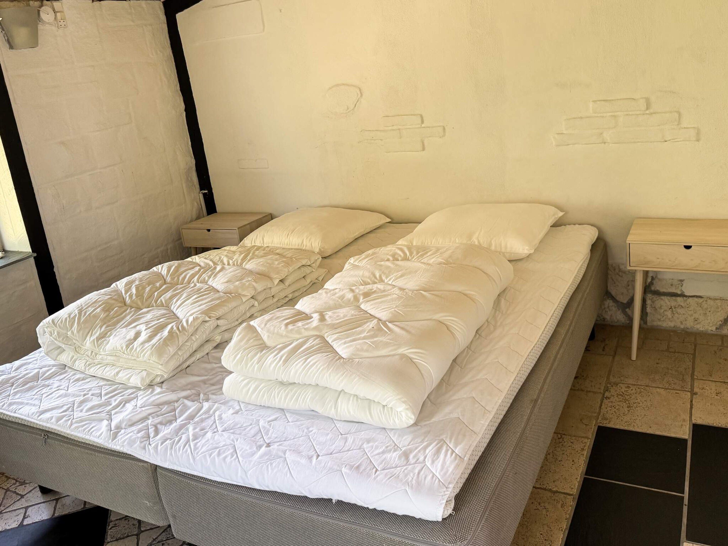A bed with a quilted mattress and two pillows, flanked by two bedside tables with drawers, in a room with a plain wall and a window.