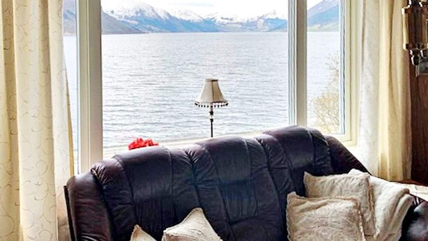 En mörkbrun lädersoffa med tre kuddar, placerad vid ett fönster med utsikt över en sjö och berg i fjärran.