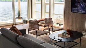 A room with a large window overlooking a lake, a sofa, two wicker chairs, a small table with a plant, and a TV mounted on the wall. - 8 Person Holiday Home in Skei i Jolster (Sunnfjord)