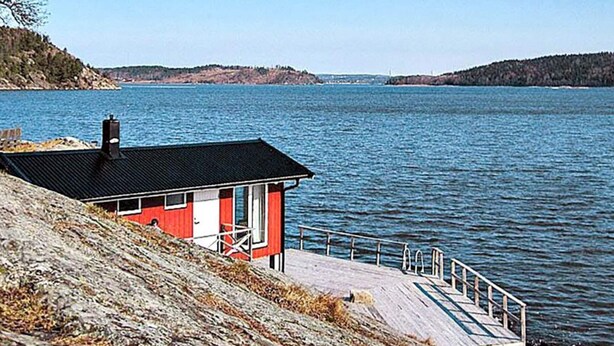 A small red building with a black roof near a body of water, a wooden dock, and rocky terrain.