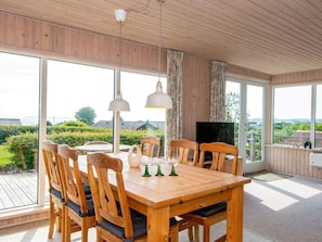 A wooden dining table with chairs, a television, and large windows offering a view of greenery and buildings.