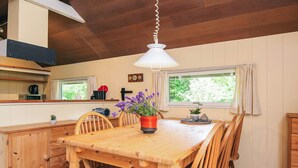 A wooden dining table with chairs, a hanging light fixture, a window with curtains, and a kitchen area with appliances and a clock on the wall. - 4 Person Holiday Home in Skjern (Skjern)