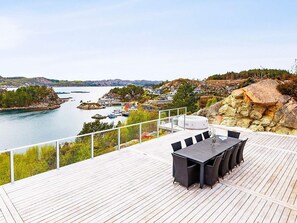 Une terrasse en bois avec une table et des chaises, donnant sur une étendue d’eau où l’on aperçoit des bateaux et des maisons au loin.