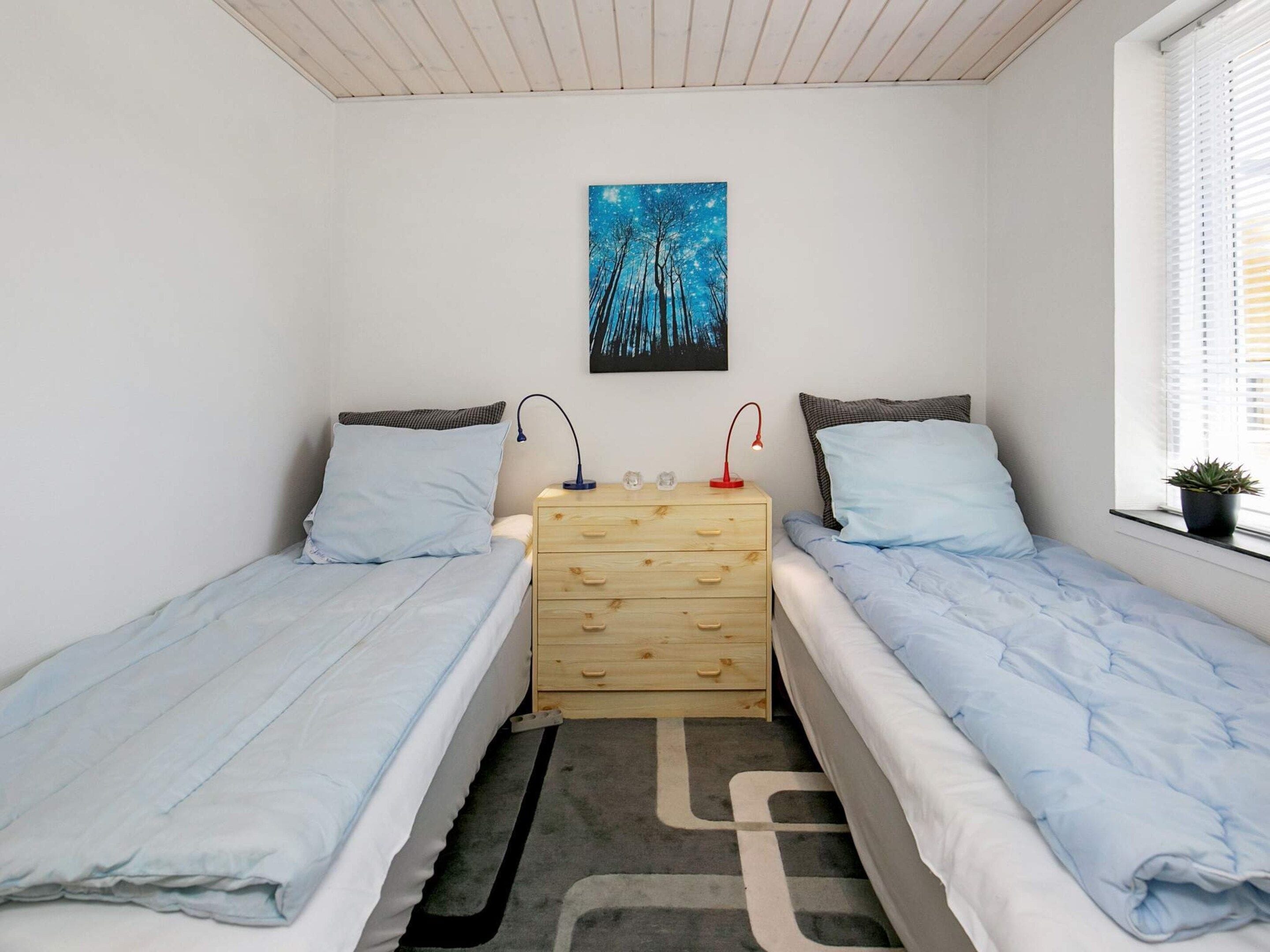 A bedroom with two single beds, a wooden dresser, a nightstand with lamps, a painting on the wall, and a window with blinds.