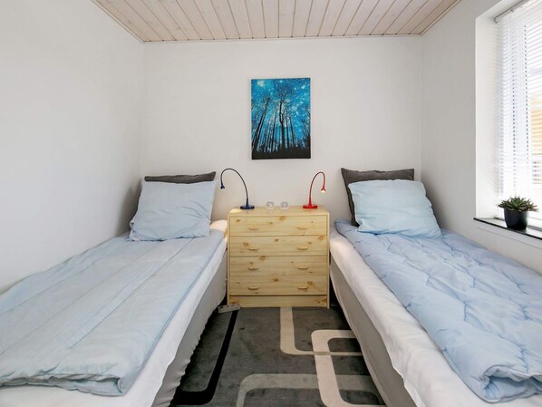 A bedroom with two single beds, a wooden dresser, a nightstand with lamps, a painting on the wall, and a window with blinds.
