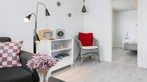 A modern living room with a sofa, a coffee table, a white bookshelf, a wicker chair with a red cushion, and a radio. - Holiday Home in LÃ¦sÃ¸ (Læsø)