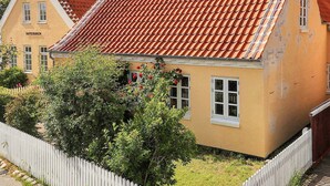 Ein gelbes Gebäude mit rotem Ziegeldach, weißem Zaun und einem Baum davor.
