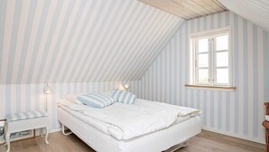 Ein Schlafzimmer mit schrägverdeckter Decke, einem Bett mit weißer Bettwäsche, einem Nachttisch, einem Stuhl und einem Fenster.