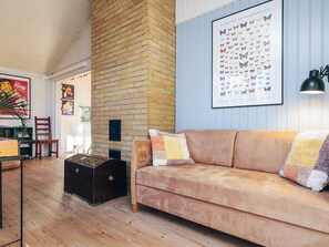 Un salon chaleureux avec un mur en briques, une grande affiche de papillons, un canapé marron et une valise ancienne.