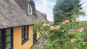 Een vakantiehuisje met rieten dak, een gele buitenkant en zwart omlijste ramen, omgeven door weelderig groen en bloeiende rozen.