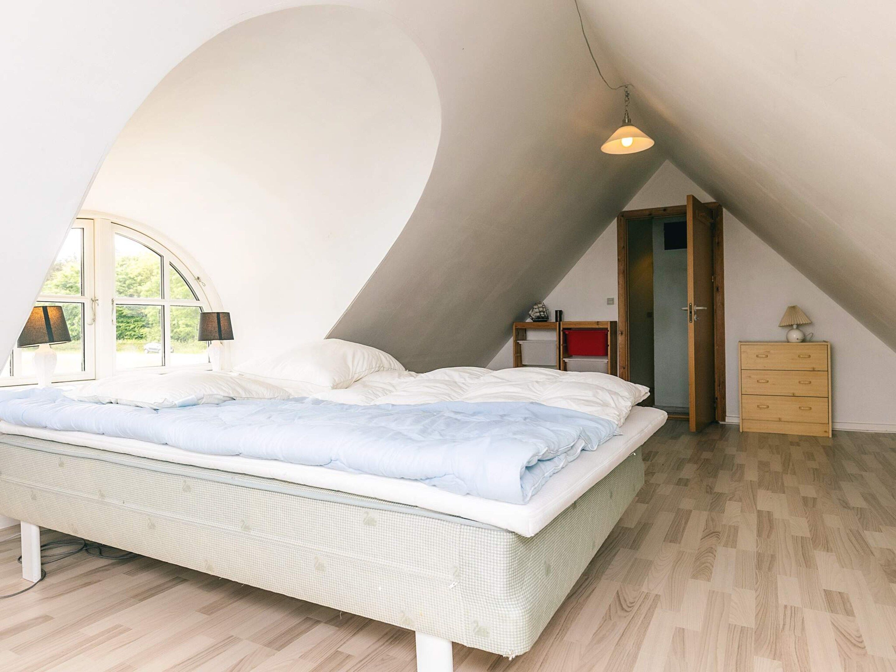 Une chambre avec un plafond en pente, un lit, une commode et une lampe.