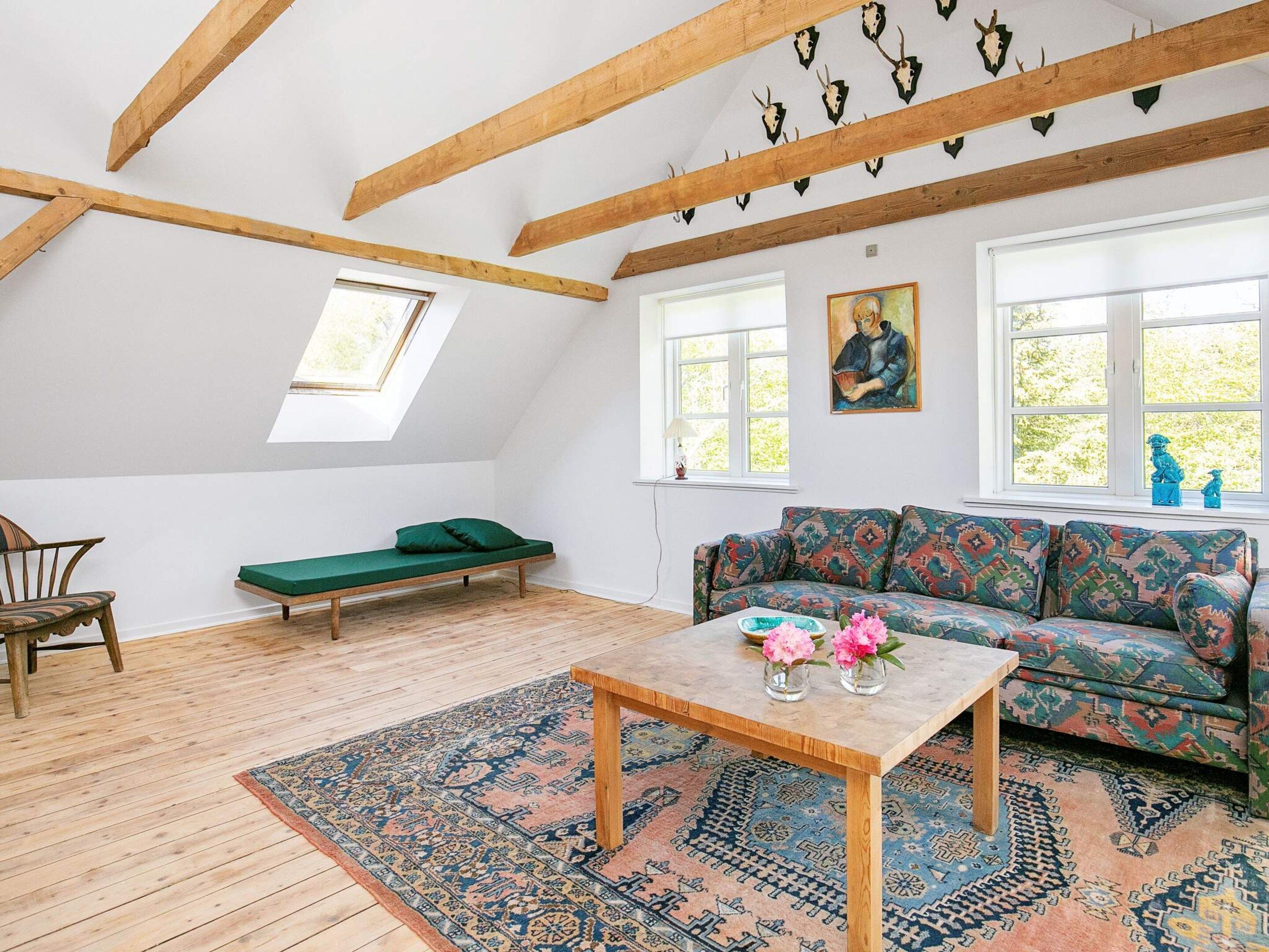 Una habitación estilo loft con techo inclinado, vigas de madera, un sofá verde, un tapete estampado y un cuadro en la pared.