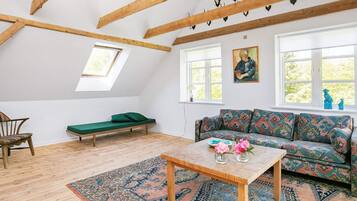 Una habitación estilo loft con techo inclinado, vigas de madera, un sofá verde, un tapete estampado y un cuadro en la pared.