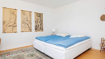A bedroom with a white bed, blue bedding, a floral rug, and three wall-mounted artworks depicting seated figures.