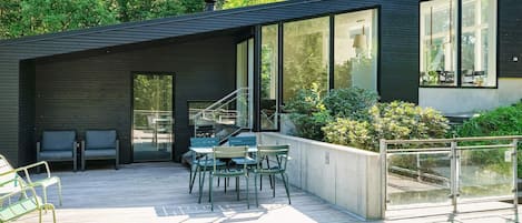 Une terrasse extérieure moderne, dotée d’une plateforme en bois, de sièges d’extérieur et de grandes fenêtres.