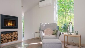 Een moderne woonkamer met een open haard, een comfortabele grijze fauteuil, een klein houten tafeltje en uitzicht op groen door grote ramen.