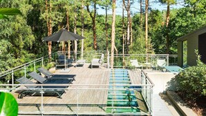 Een houten terras met een zwembad met glazen bodem, lounge stoelen en een kleine tafel.